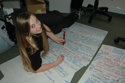 Colour photograph of a young woman lying on the floor surrounded by sheets of paper, writing lists on them.