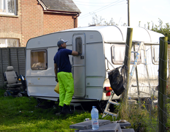 Colour photograph of a man in fluorescent plastic trousers, boots and a baseball cap, cleaning the outside of a white caravan.