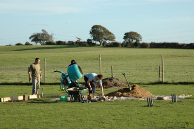 Colour photograph of young men laying foundations on a grassy site, with a grassy field behind them