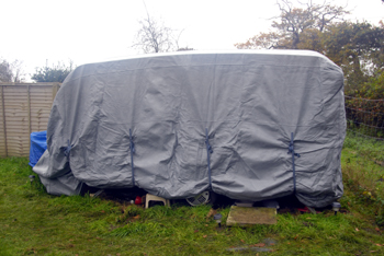 Colour photograph of a caravan shape under a grey cover, with white sky behind.