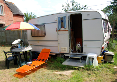 Colour photograph of the caravan with Genie standing on the steps, and an orange sunbed and table and chairs with sun umbrella up in the foreground.