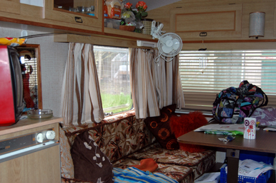 Colour photograph of the inside of a caravan, with contents including a red television and pub mirror, orange and brown cushions and a vase of orange roses.