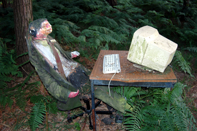 Colour photograph of a papier mache figure of a man, dressed in a suit and sitting behind a desk with a computer on it. He is surrounded by ferns.
