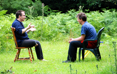 Colour photograph of two white men, sitting on chairs facing each other with ferns and woodland behind them. David is signing and speaking on the left, while Colin listens on the right.