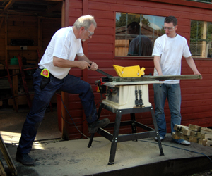 Colour photograph of two men working at a saw horse, with a shed in the background.