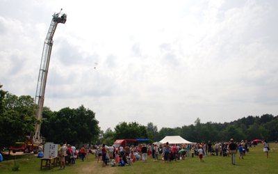 Colour photograph of the fire engine ladder rising up over the summer fair, with a bear parachuting down.