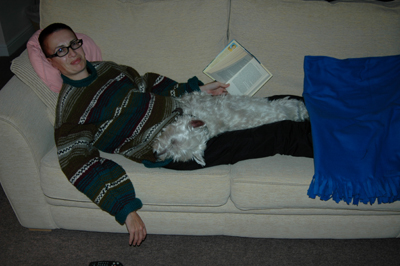 Colour photograph of woman lying on a sofa holding a book, with a dog lying on top of her