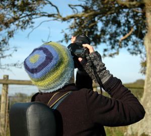 Close up colour photograph, taken from behind, of me in a black fleece and blue and green knitted hat, taking a photograph of a tree.