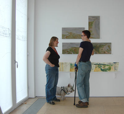 Colour photograph of Sally and I standing talking in front of oil paintings of the Holton Lee foreshore, with Genie waiting patiently