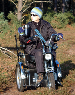 Colour photograph of me posing on my off-road scooter at the edge of the woods, thermos mug in hand. I am wearing a black fleece, black nylon trousers and jacket, boots and a blue and green stripy hat and gloves, with black sunglasses too.