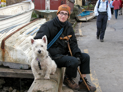 Colour photograph of myself and Genie, sitting on a wall beside the road and looking towards the camera, with boats laying upside down in the background.