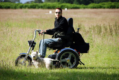 Colour photograph of the artist on a black and chrome three-wheeled off-road mobility scooter, dog beside her