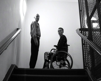 Black and white video still of two people looking down from the top of a staircase - a woman using a manual wheelchair, facing a man who is standing.