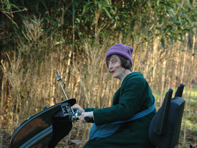 Colour photograph of the top half of a woman (in profile) dressed in a green coat with a purple ski hat and blue bag, riding an off-road scooter with woodland in the background