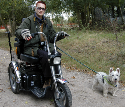 Colour photograph of the artist on an outdoor scooter, dressed for the cold weather, with Genie the Westie on a lead.