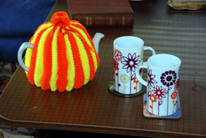 Close-up colour photograph of a teapot, covered by an orange and yellow teacosy, with two floral mugs.
