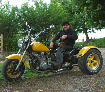 Colour photograph of a smiling man sitting on a large three-wheeled yellow motorbike in a lane in front of trees and fencing.
