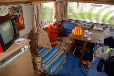 Colour photograph of the inside of the caravan, including a red portable television, blue fluffy rug, orange and brown furnishings and so on.