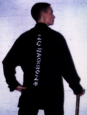 Photograph of Ju standing with her walking stick with her back to the viewer, wearing a black shirt with the words "No Backbone" printed down her spine