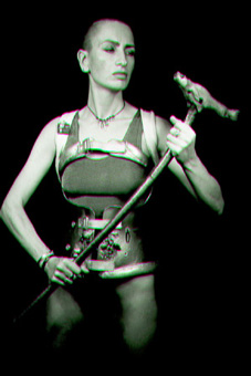Black and white photo showing ju90, dressed in her decorated spinal brace, a silver vest and black g-string, standing holding her walking stick in front of her like a weapon and looking down at it.