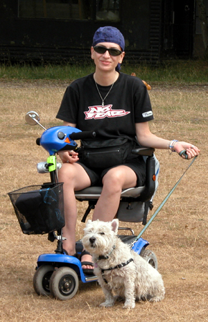 Colour photograph of me sitting smiling on my blue mobility scooter, wearing black sunglasses, a purple head scarf, and black shorts and a black T shirt with a 'No Fear' logo. Genie is straining at her lead.