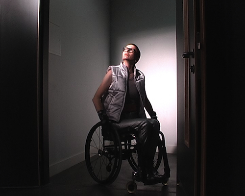 Atmospheric photograph of Ju dressed in silver, with bare arms, black framed glasses and a minimalist black wheelchair, glimpsed through a doorway looking up into light.